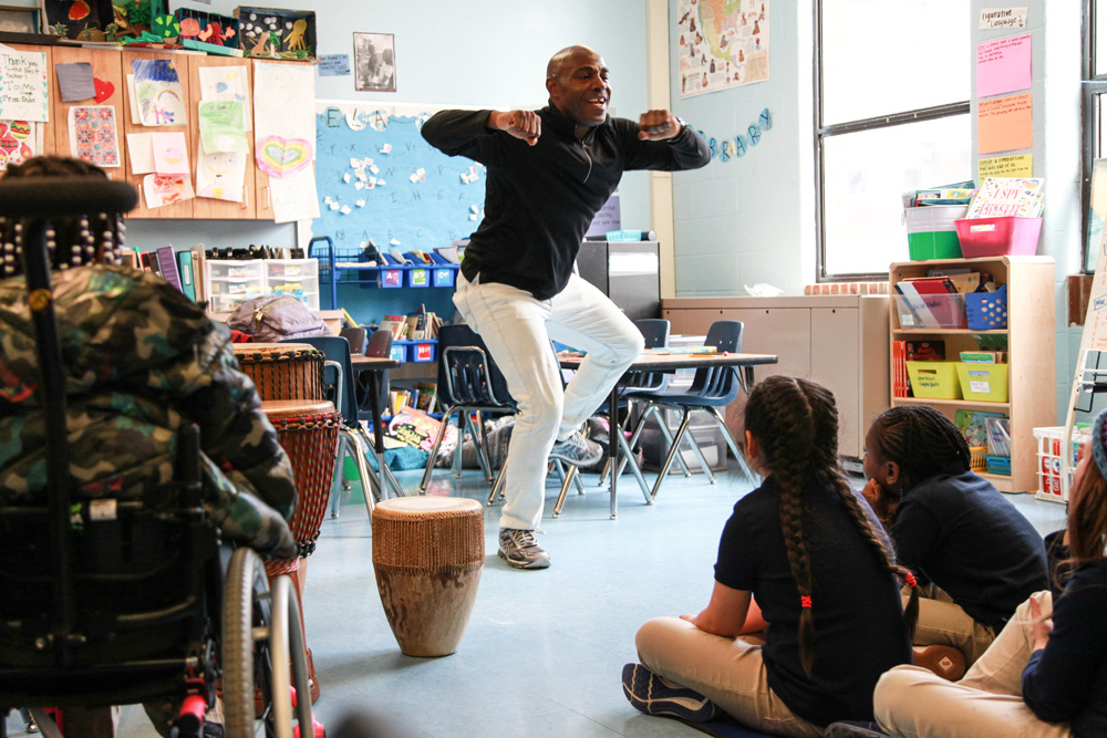 Teaching artist Ssuuna dances as children seated around him watch intently