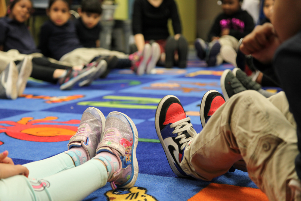 Students and teachers sit in a circle on the floor, feet outstretched. Children imagine their colorful sneakers are transformed into tap shoes and are in position to stomp out beats.