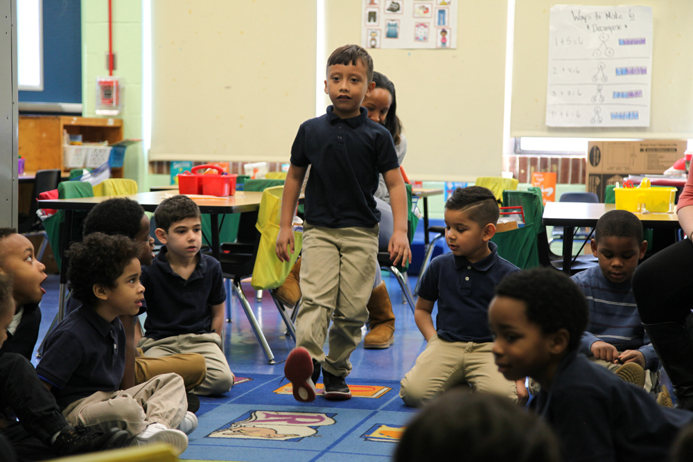 A kindergarten student models a "heel-toe" tap movement by striking the heel of his sneaker to the floor. His classmates sit in a circle on the rug and watch carefully.