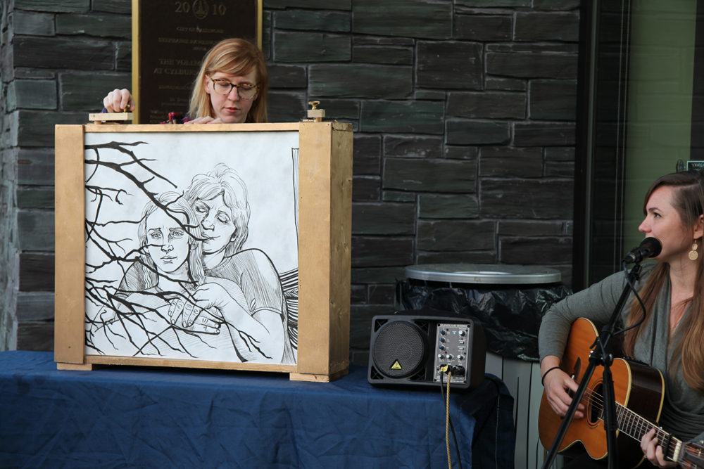 Artist Maura Dwyer turns a large scroll revealing a hand-drawn illustration of woman with her arms enveloping a younger woman's shoulders. Playing guitar beside the artist is folk musician Letitia VanSant.