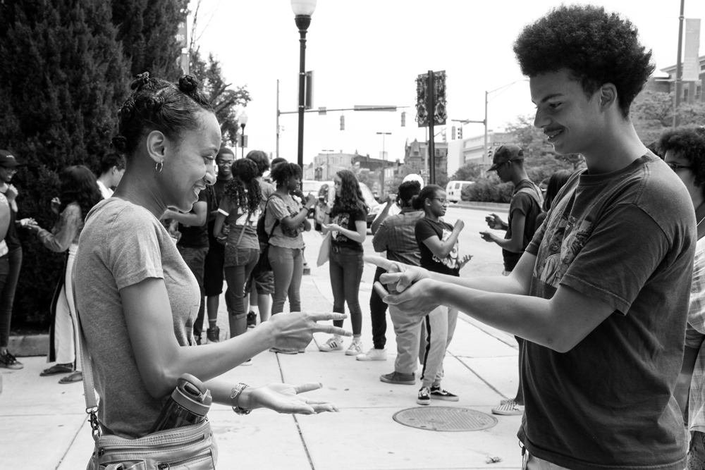 Two high school interns playing Rock, Paper, Scissors on the sidewalk outside of Motor House on North Ave. in Baltimore. A larger group of high school students are gathered in the background.