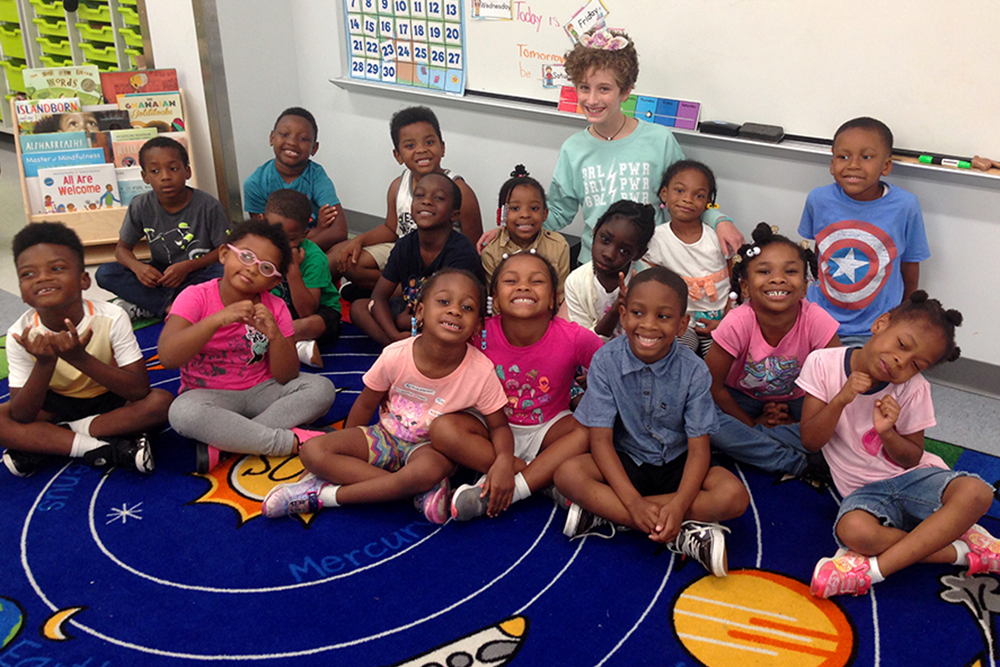 The Pre-K class sitting together on a rug depicting the solar system, posing with Alice, a student volunteer.