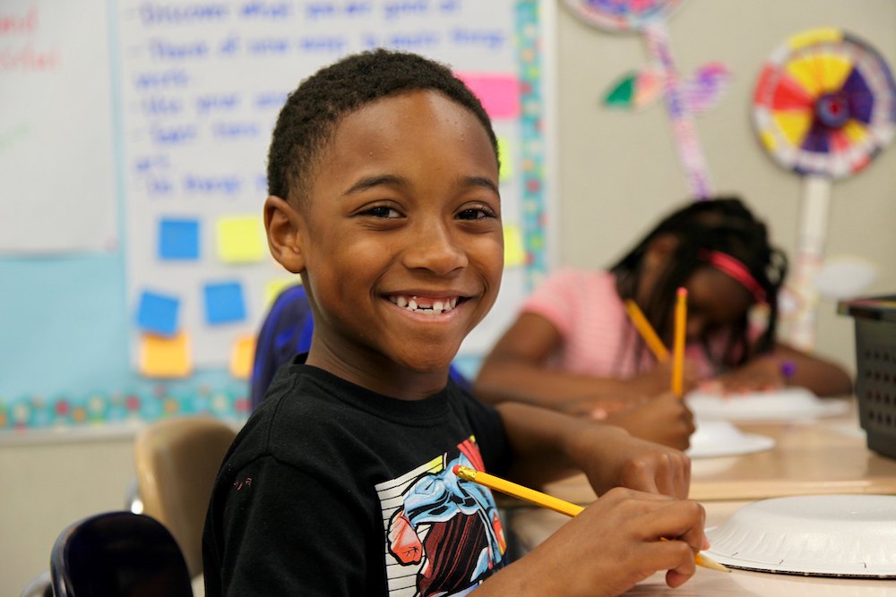 Student sitting at a desk, pencil in hand, looking into the camera and smiling.