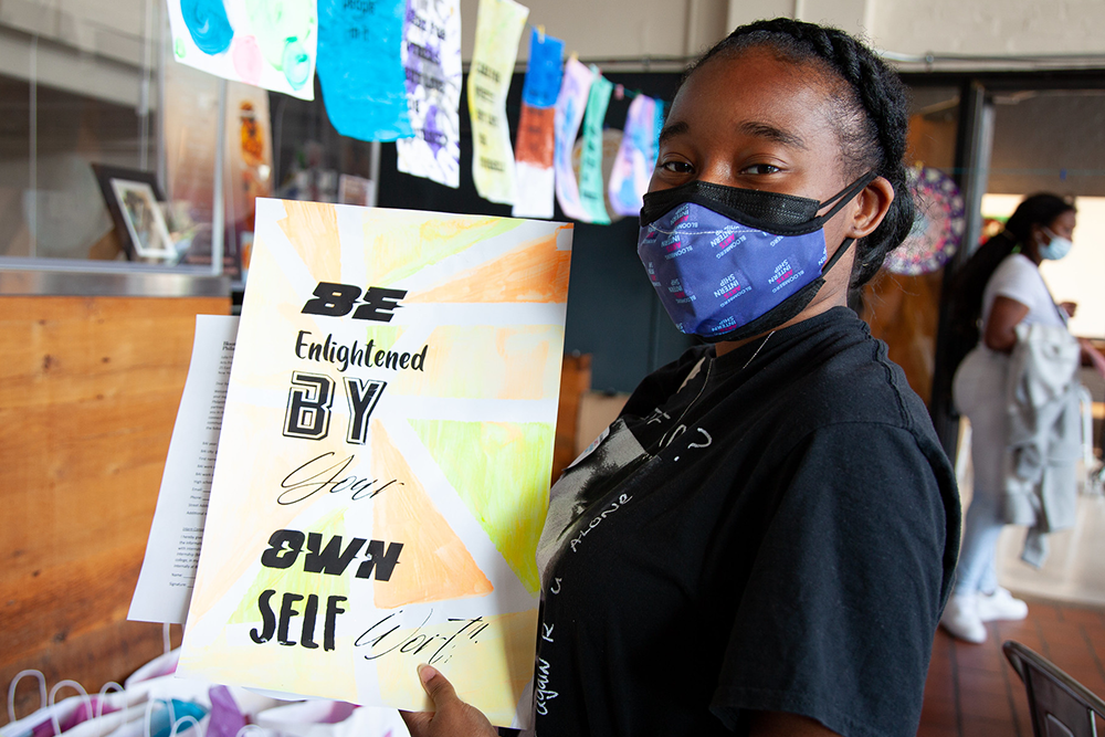 A student in a mask holds up a poster that says, "Be enlightened by your own self worth."