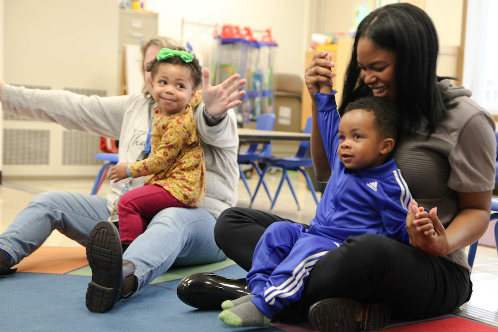 Two toddlers, one with a green bow in their hair and the other in a blue tracksuit, sitting on the laps of two adults on the floor of a classroom.