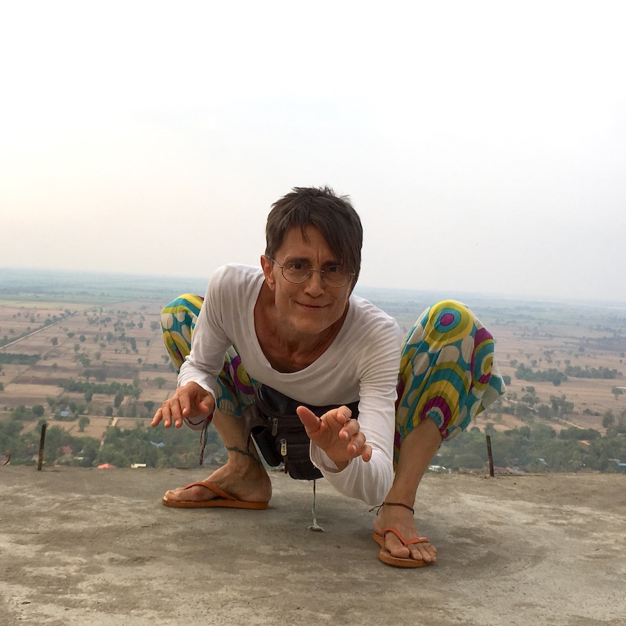 Woman in brightly patterned pants crouching on land overlooking farmland.