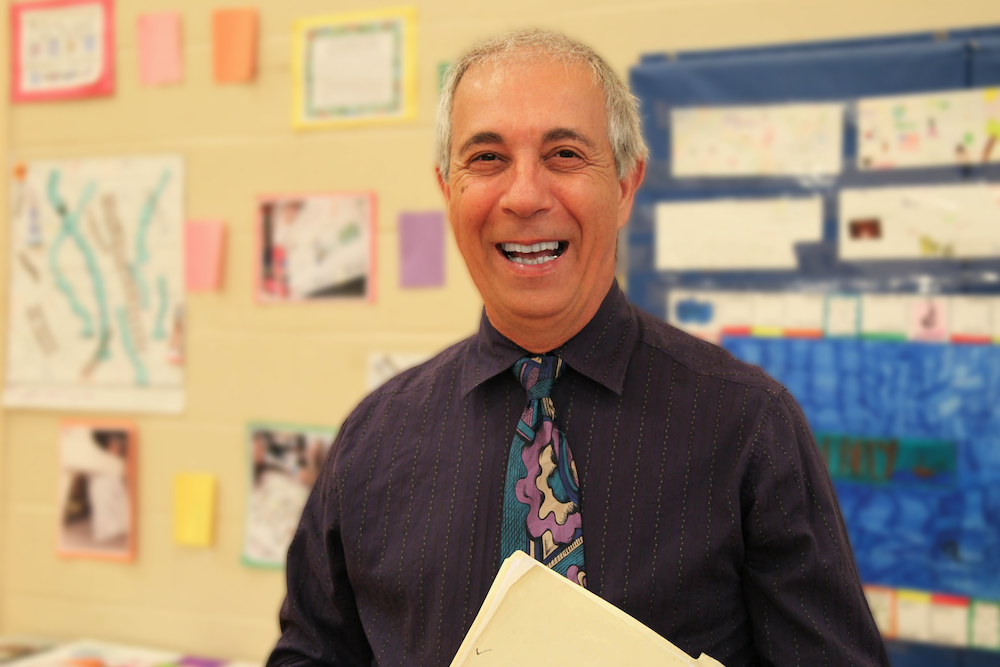 White man with short gray hair with a bright smile standing in front of a school bulletin board and holding a manilla folder.