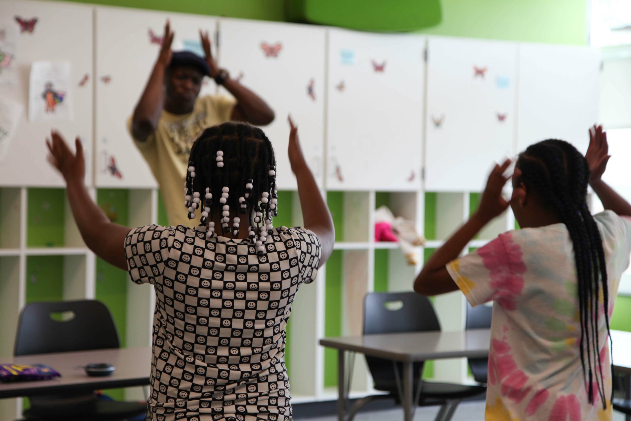 Femi the Drifish stands in front of students in a lime green classroom lined with white cabinets. The artist's hands are held near his head and the students are imitating his gestures.