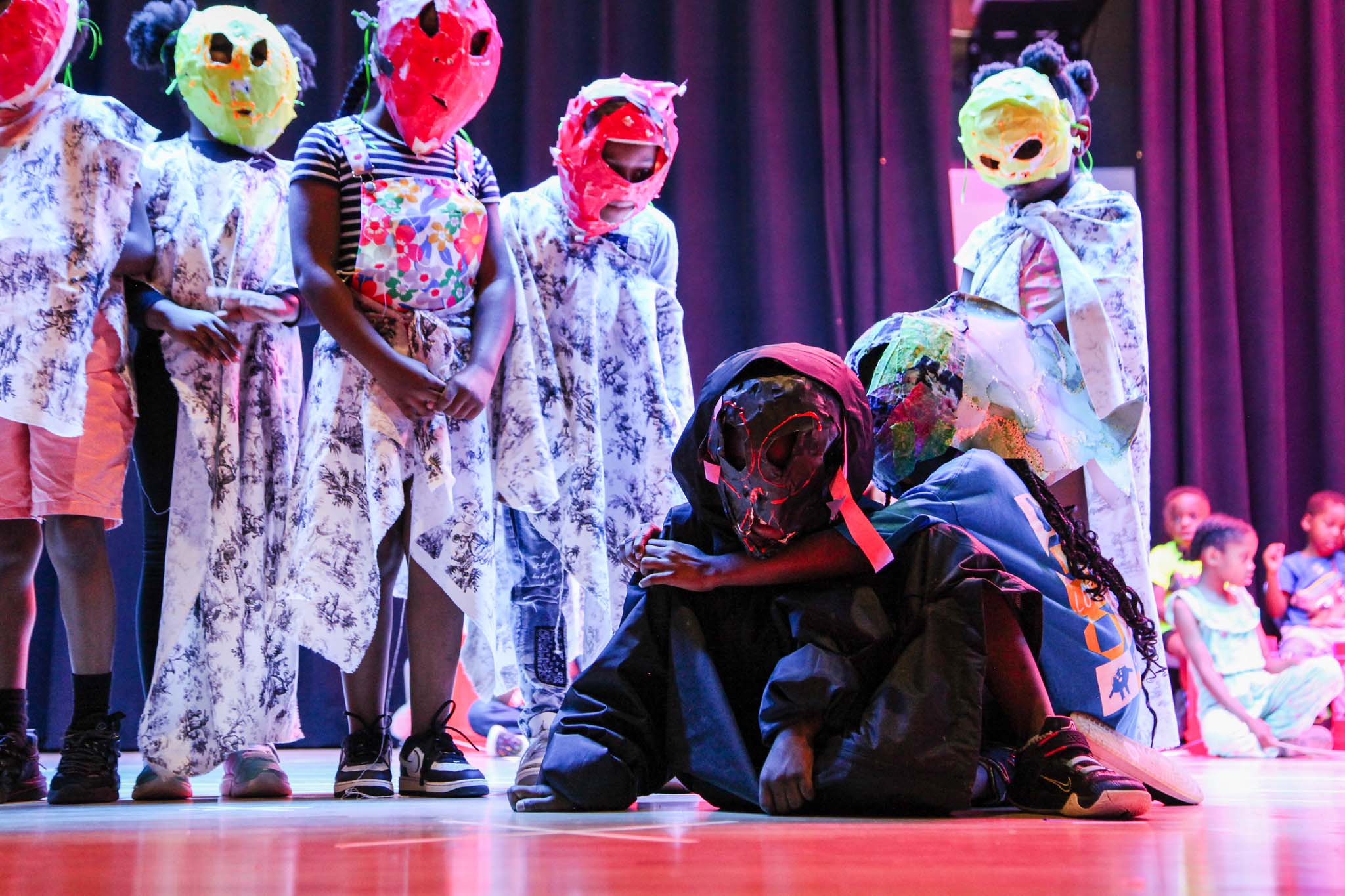 Children are draped in patterned fabric and wearing paper maché masks on a stage. Most are standing, but two are huddled on the ground. One is draped in all black fabric and wearing a black and red mask.