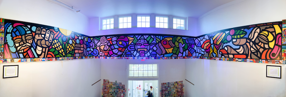 A mural with lines and colors like stained glass and the Mayan calendar at the center runs on three walls of a school's entrance hall above the doors. windows line the top of the mural.